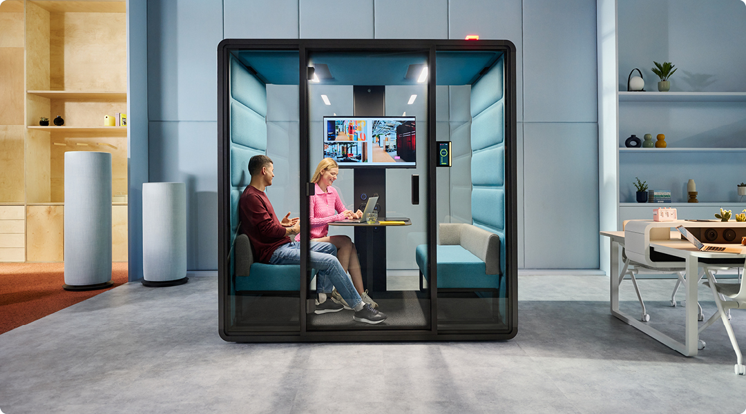 Two people collaborating inside a modern, glass-enclosed acoustic meeting pod with padded interiors, a screen display, and ergonomic seating, set within a stylish and minimal office space.