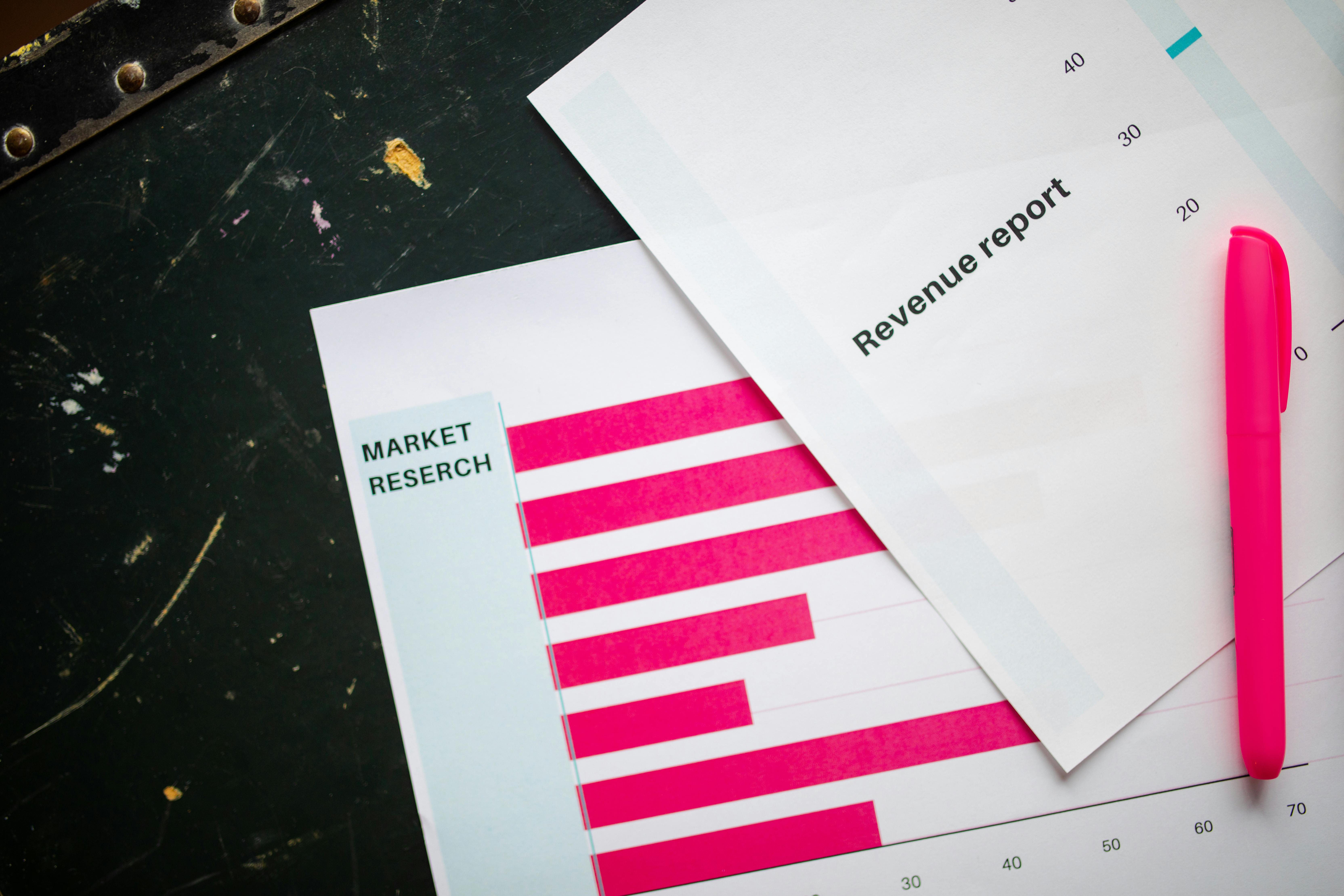 Close-up of business documents showing a revenue report and a market research chart with a pink bar graph, placed on a desk with a pink pen.