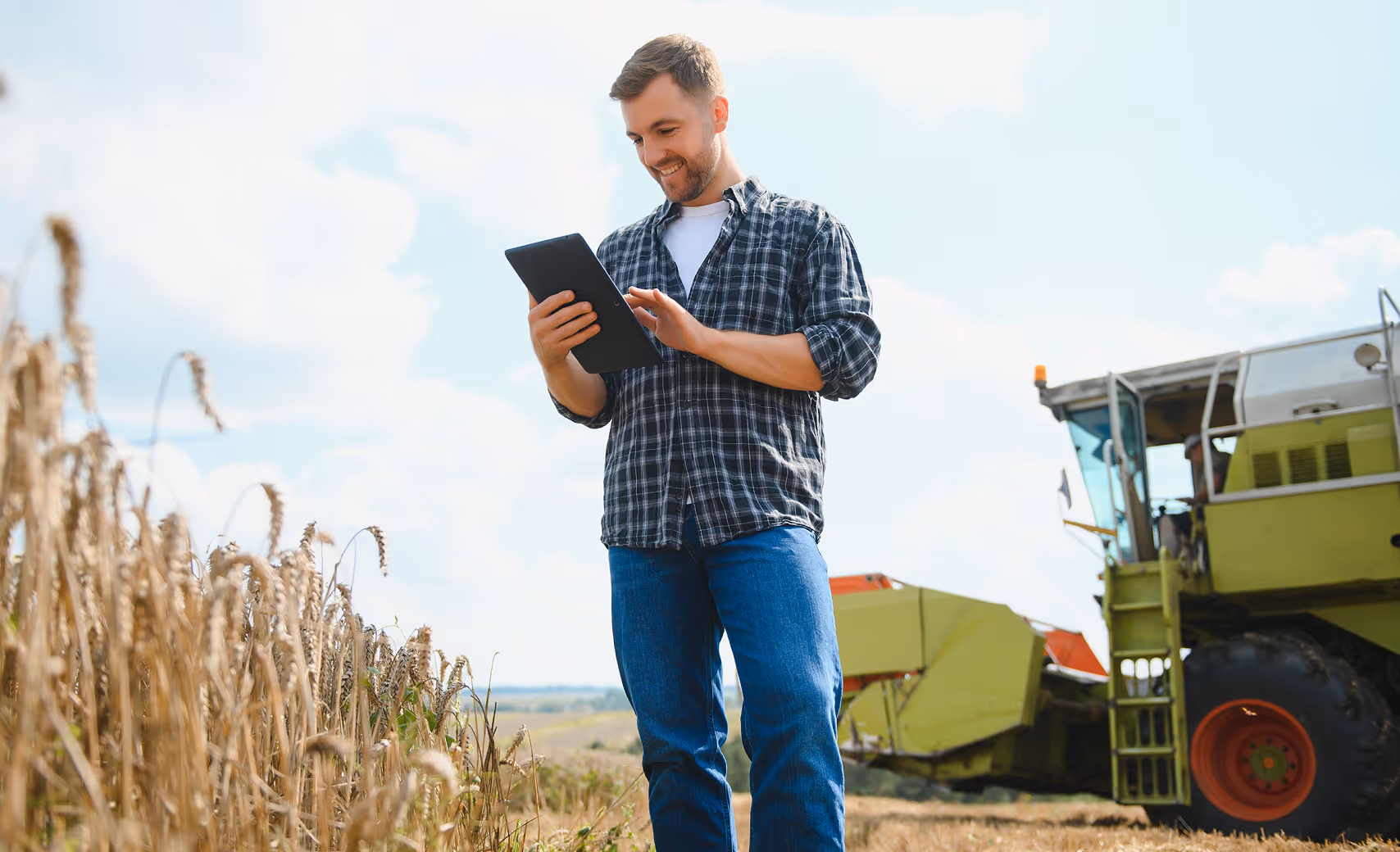Smiling farmer standing in wheat field using a tablet with a combine harvester in the background.