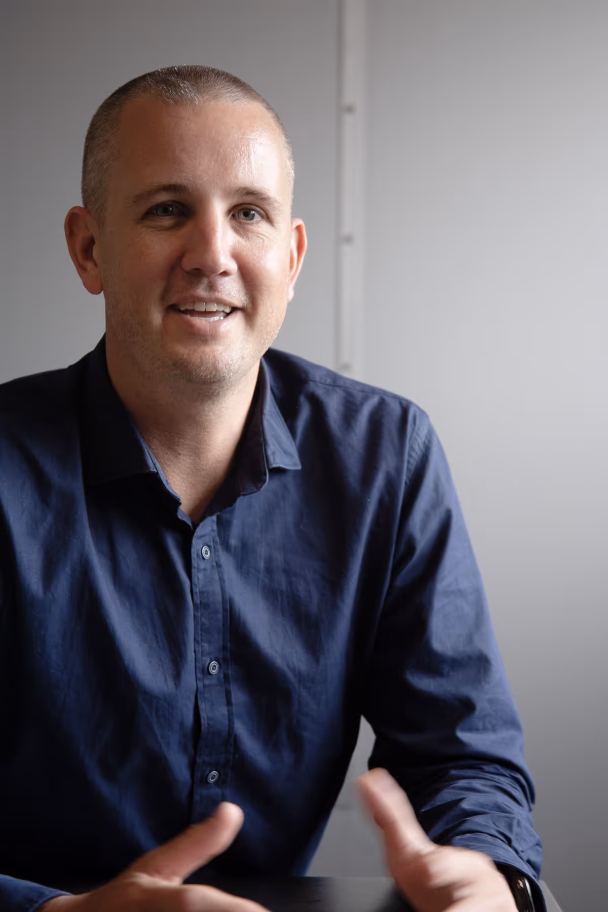 Man in a dark blue shirt sitting at a table, speaking with hands gesturing.