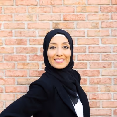 Woman smiling and wearing a black hijab and black jacket standing in front of a brick wall.