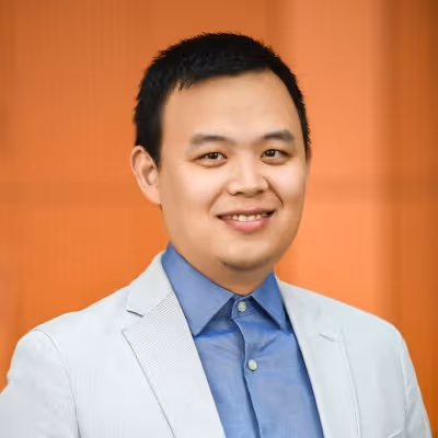 Smiling man with short black hair wearing a light gray blazer and blue shirt against an orange background.