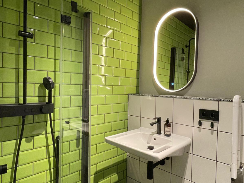 Modernes Badezimmer mit glänzenden grünen Metrofliesen an der Wand, weißen quadratischen Fliesen um ein wandmontiertes Waschbecken mit schwarzem Wasserhahn, hinterleuchtetem ovalen Spiegel und schwarzen Duscharmaturen hinter einer Glastür.