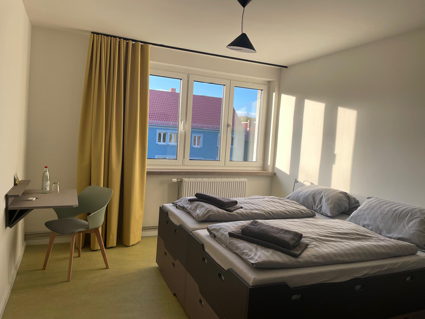 Minimalistisches Gästezimmer mit zwei zusammengestellten Einzelbetten, grauer Bettwäsche, gelbem Vorhang, kleinem Schreibtisch mit Stuhl und Tageslicht, das durch das Fenster fällt.