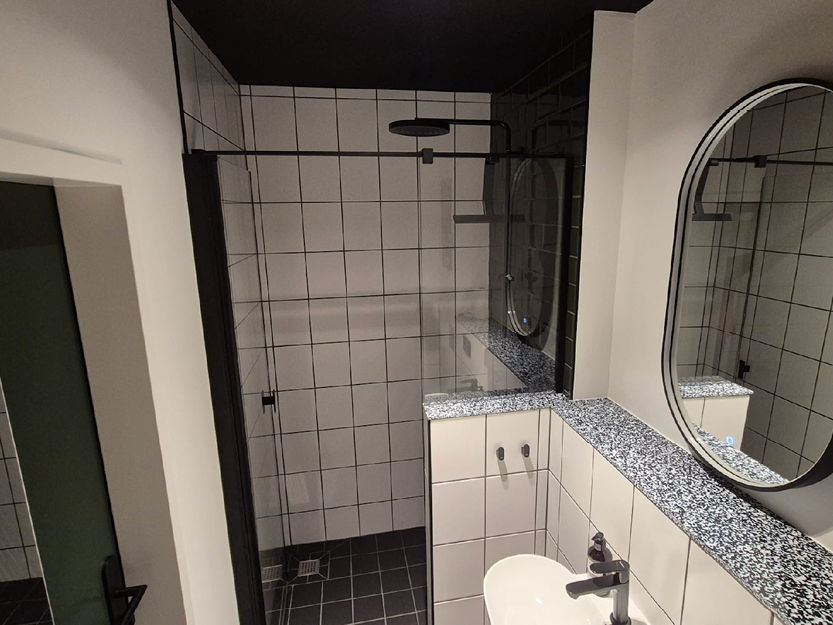 Modernes Badezimmer mit schwarzem Fliesenboden, weißen Fliesenwänden, einer verglasen Duschkabine mit Regenduschkopf, einem weißen Waschbecken und einem ovalen Spiegel über einer gesprenkelten Arbeitsplatte.