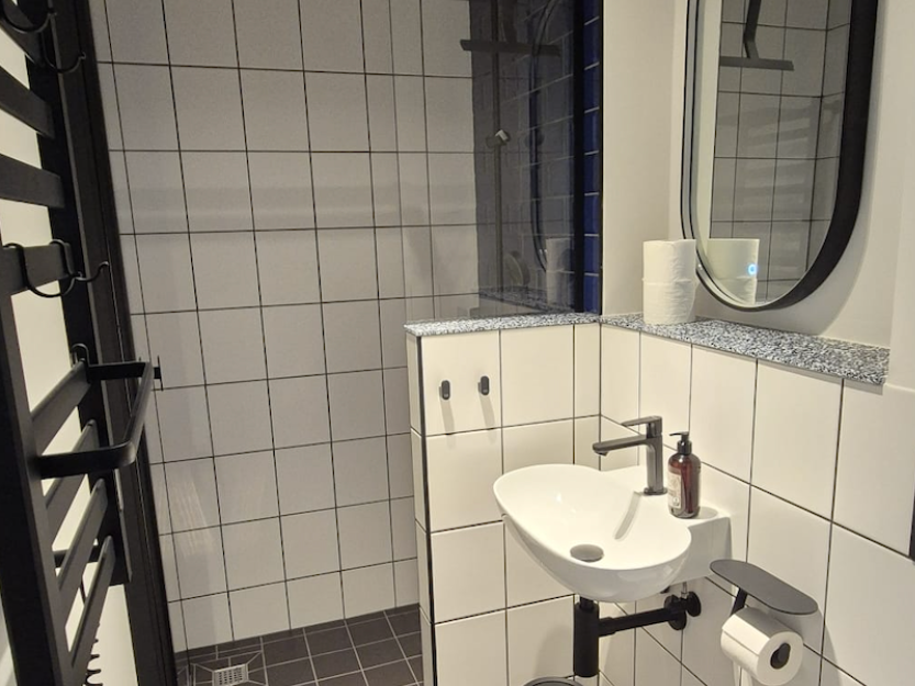 Minimalistisches Badezimmer mit weißen Fliesen an Wänden und Boden, einem weißen Waschbecken, einem Spiegel und einem schwarzen Handtuchhalter.