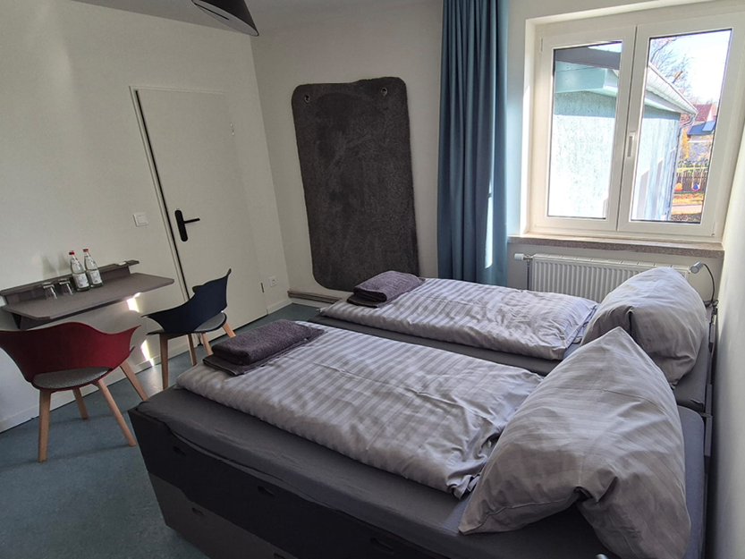 Kleines Gästezimmer mit zwei zusammengestellten Einzelbetten, jedes mit grau gestreifter Bettwäsche und einem gefalteten Handtuch darauf, neben einem Fenster mit blauen Vorhängen und zwei Stühlen neben einem Wandregal.