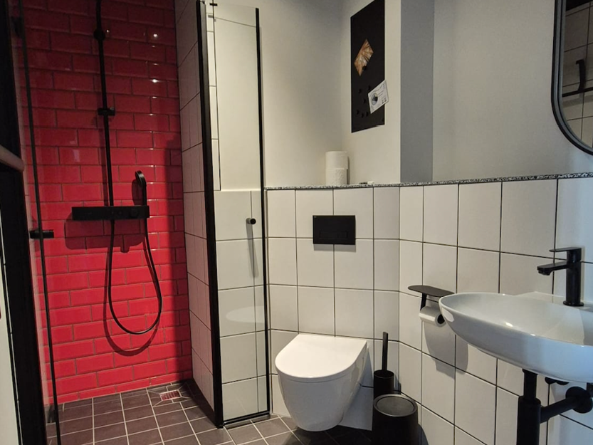 Modernes Badezimmer mit weiß gefliesten Wänden, einer wandhängenden Toilette, schwarzen Armaturen, einem weißen Waschbecken und einem Duschbereich mit roten Fliesen im Ziegelsteinmuster.