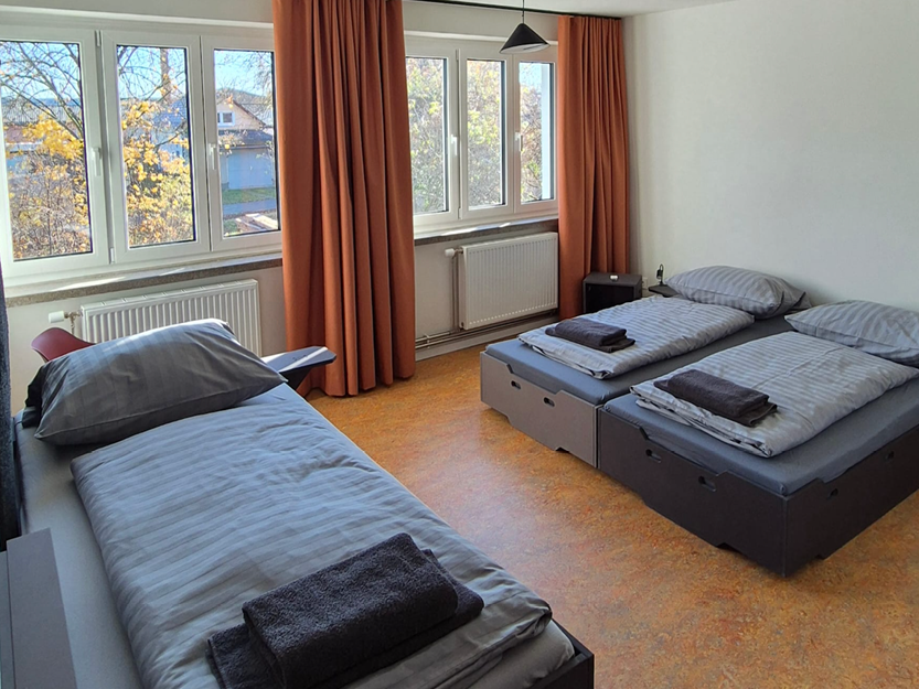 Minimalistisches Gästezimmer mit drei Einzelbetten, jedes mit grau gestreifter Bettwäsche und gefalteten dunklen Handtüchern, großen Fenstern mit orangefarbenen Vorhängen und einem hellbraun-orangenem Boden.