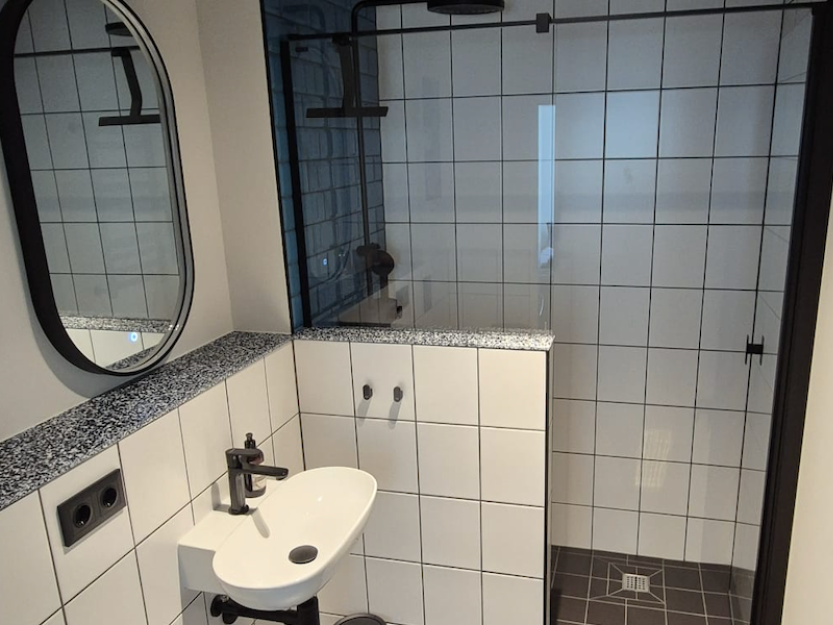 Modernes Badezimmer mit weiß gefliesten Wänden, kleinem weißen Waschbecken mit schwarzem Wasserhahn, rundem Spiegel und verglaster Dusche mit dunklen Bodenfliesen.