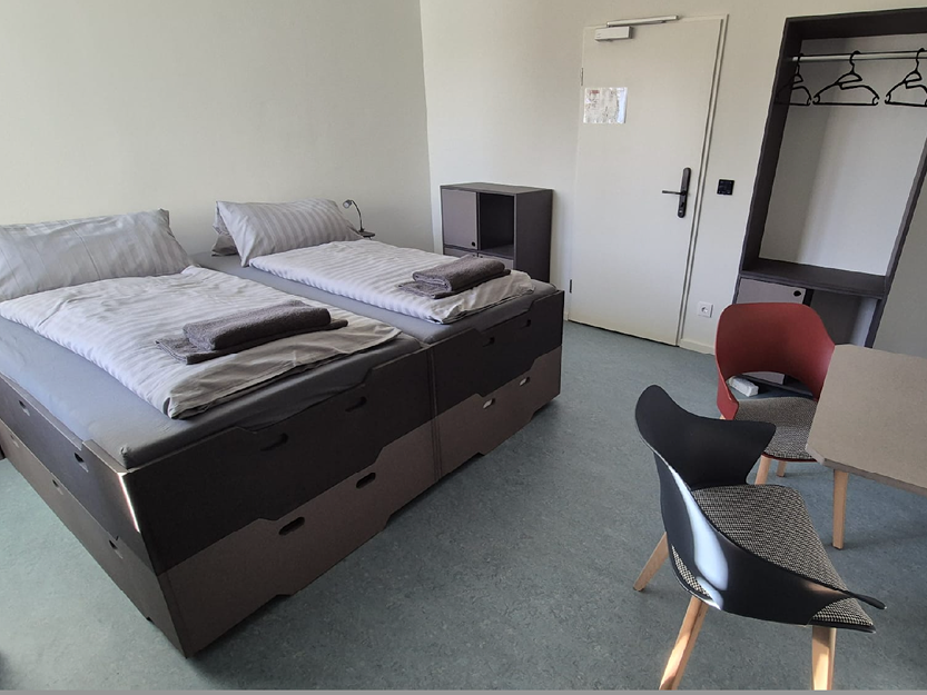 Minimalistisches Gästezimmer mit zwei zusammengestellten Einzelbetten, grauer Bettwäsche, Handtüchern darauf, zwei modernen Stühlen, einem kleinen Tisch und einem offenen Kleiderschrank mit Kleiderbügeln.