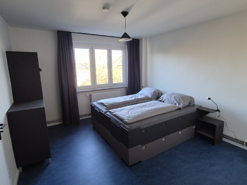 Minimalistisches Gästezimmer mit zwei zusammengestellten Einzelbetten, grauer Bettwäsche, einem Fenster mit dunklen Vorhängen, einer Pendelleuchte und schlichten dunklen Möbeln, darunter ein Schrank und ein Nachttisch.