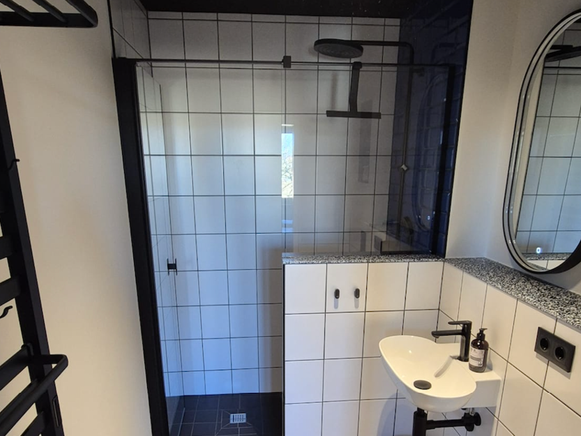 Modernes Badezimmer mit weiß gefliesten Wänden und schwarzen Fugen, mit einer verglasen Dusche mit schwarzen Armaturen, einem kleinen weißen Wandwaschbecken, einem runden Spiegel und einem schwarzen Handtuchhalter.