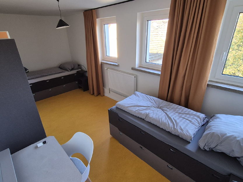 Einfaches, aufgeräumtes Zimmer mit zwei Einzelbetten, weißen Wänden, orange-gelbem Boden, dunkelgelben Vorhängen und einem weißen Stuhl an einem kleinen Schreibtisch.