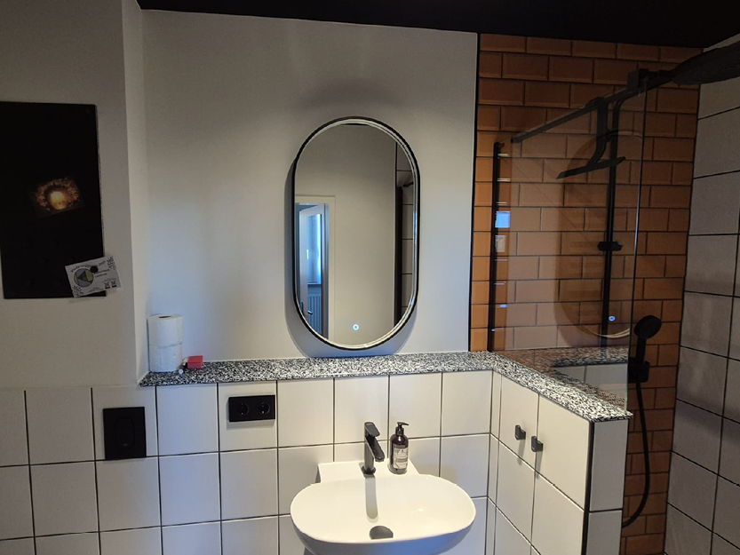 Moderne Badezimmerecke mit weiß gefliesten Wänden, Granitarbeitsplatte, ovalem Spiegel, weißem Waschbecken, schwarzem Wasserhahn, Seifenspender und einer verglasen Dusche mit orangefarbenen Fliesen.