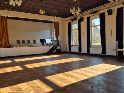 Leere Veranstaltungshalle mit Holzparkettboden, Bühne mit Stühlen und Klavier, große Fenster, durch die Sonnenlicht hereinfällt.