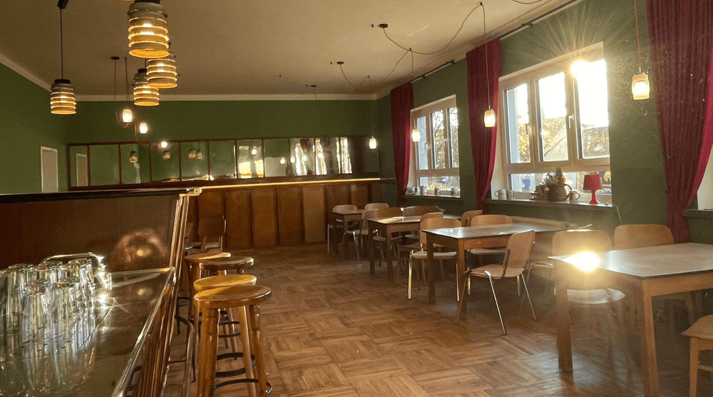 Sonnendurchfluteter Saal mit Holztischen und -stühlen entlang grüner Wände mit roten Vorhängen und hängenden Leuchten.