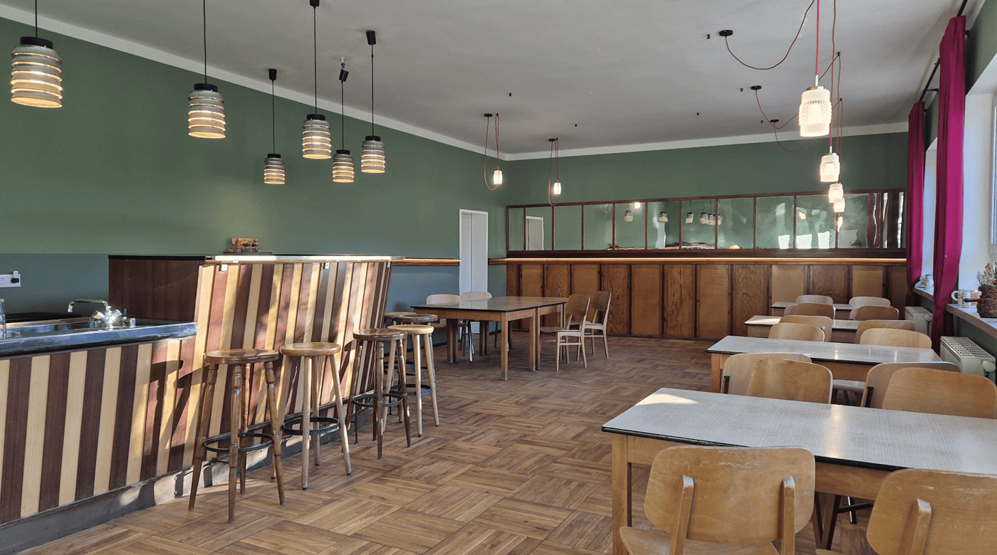 Leeres Café/Bar-Interieur mit Holzhockern an einer gestreiften Bar, quadratischen Tischen, Holzstühlen, grünen Wänden, hängenden Leuchten, roten Vorhängen und Parkettboden.