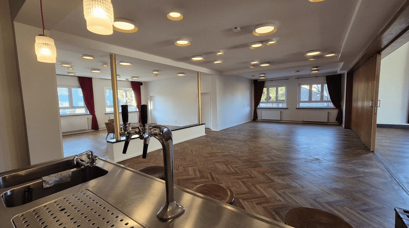 Innenansicht eines großzügigen Barbereichs mit Fischgrätenparkett, mehreren Einbauleuchten an der Decke, roten Vorhängen an den Fenstern und einer Bierzapfanlage aus Edelstahl im Vordergrund.