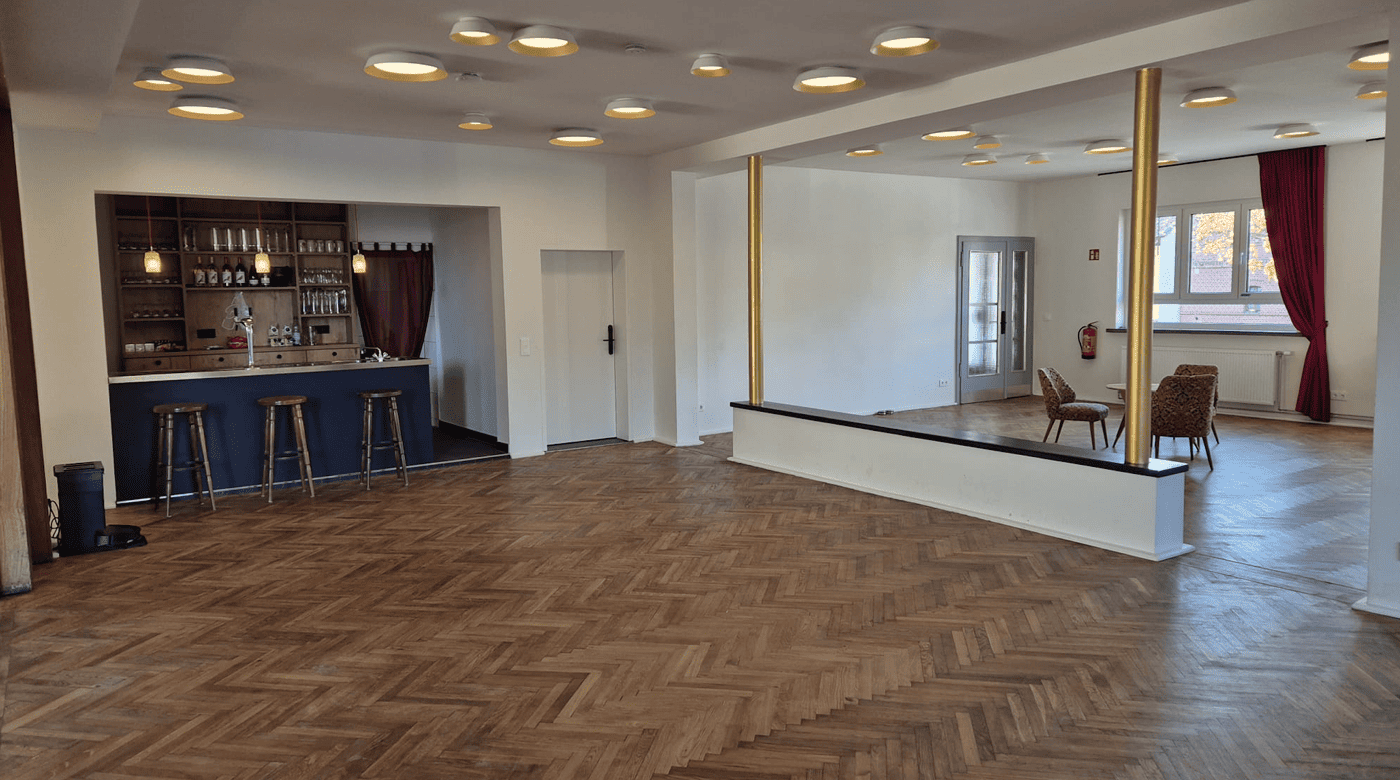 Geräumiger Raum mit Fischgrätenparkett, einer kleinen Bar mit drei Hockern auf der linken Seite, zwei goldenen Säulen und zwei Sesseln in der Nähe eines Fensters mit roten Vorhängen auf der rechten Seite.