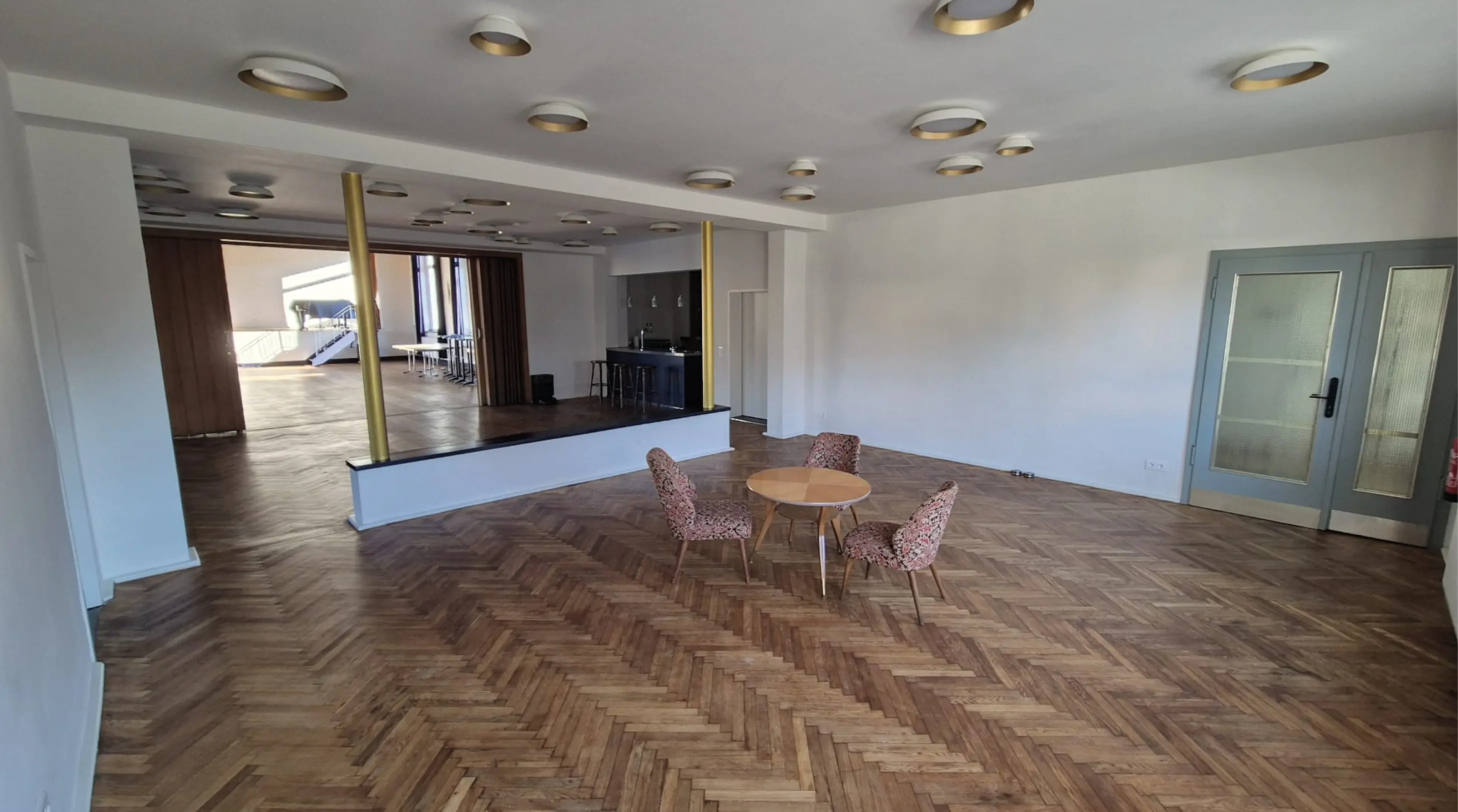 Geräumiger Raum mit Fischgrätparkett, drei Stühlen mit Blumenmuster um einen runden Holztisch und einem kleinen Barbereich im Hintergrund.