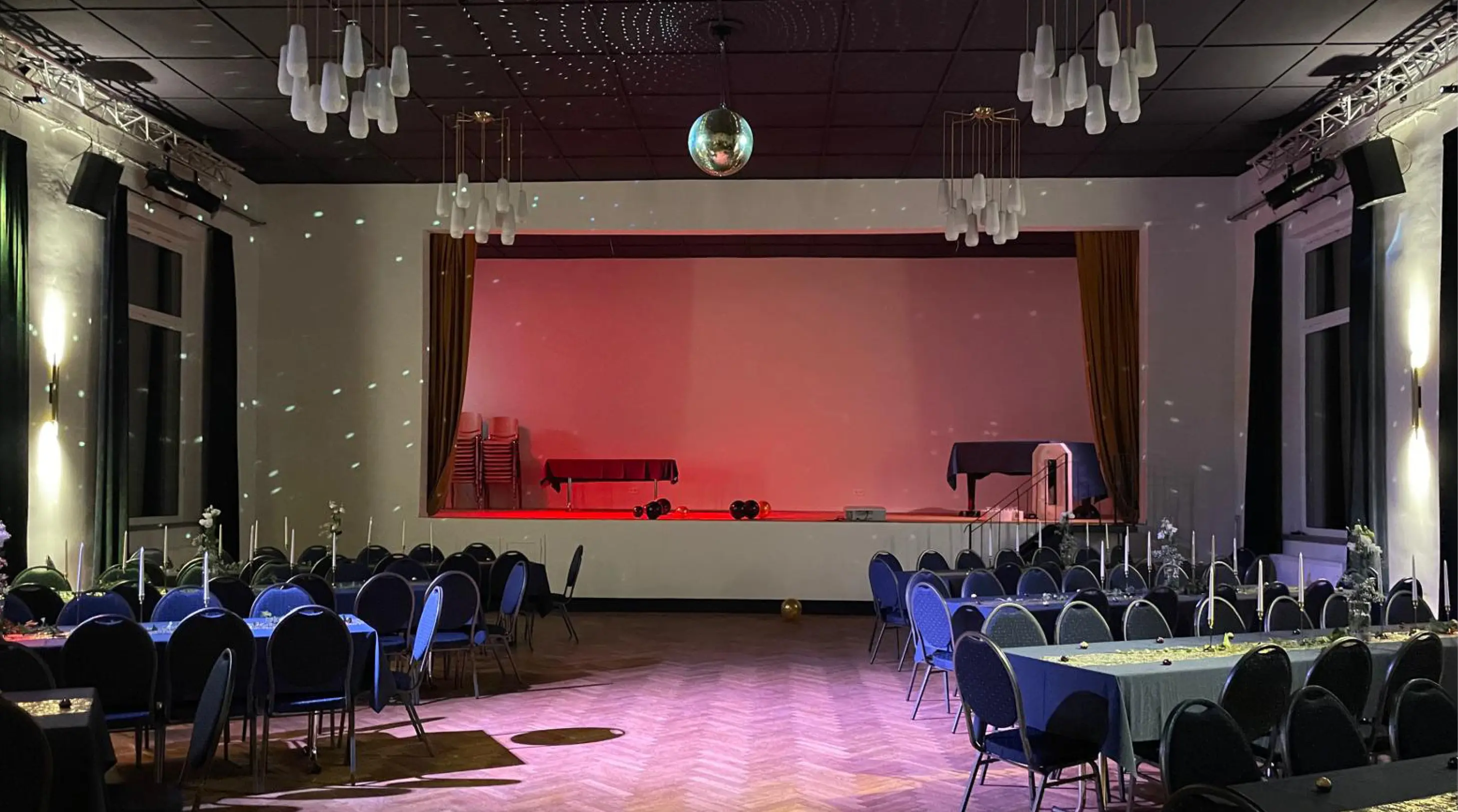 Ein großer Veranstaltungssaal mit ordentlich angeordneten Tischen und Stühlen, einer kleinen Bühne, die mit rotem Licht beleuchtet und mit einer Discokugel dekoriert ist.