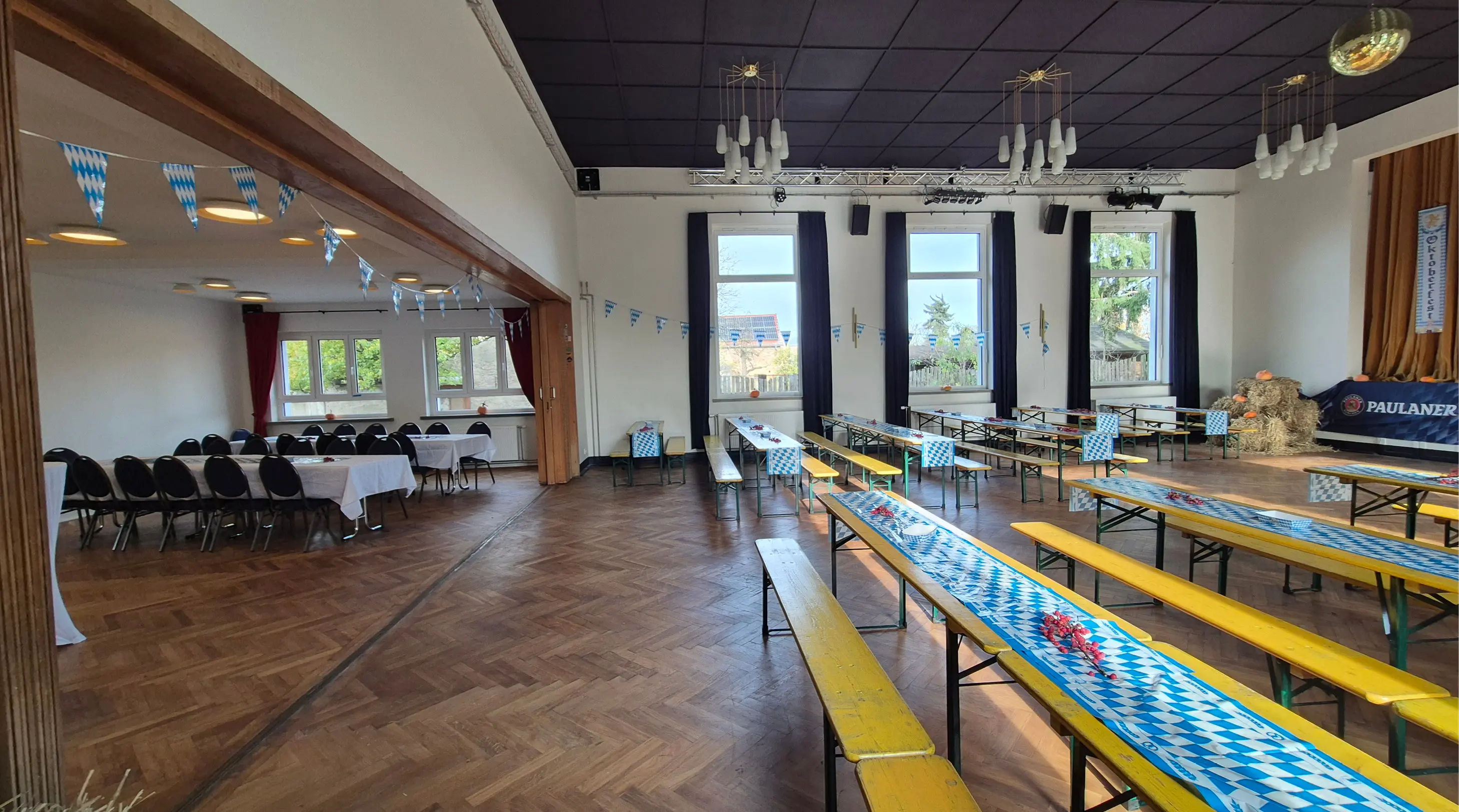 Großer Veranstaltungssaal mit gelben Holzbänken und Tischen, die mit blau-weißen Tischläufern und Flaggen im bayerischen Stil dekoriert sind, sowie Heuballen in der Ecke.
