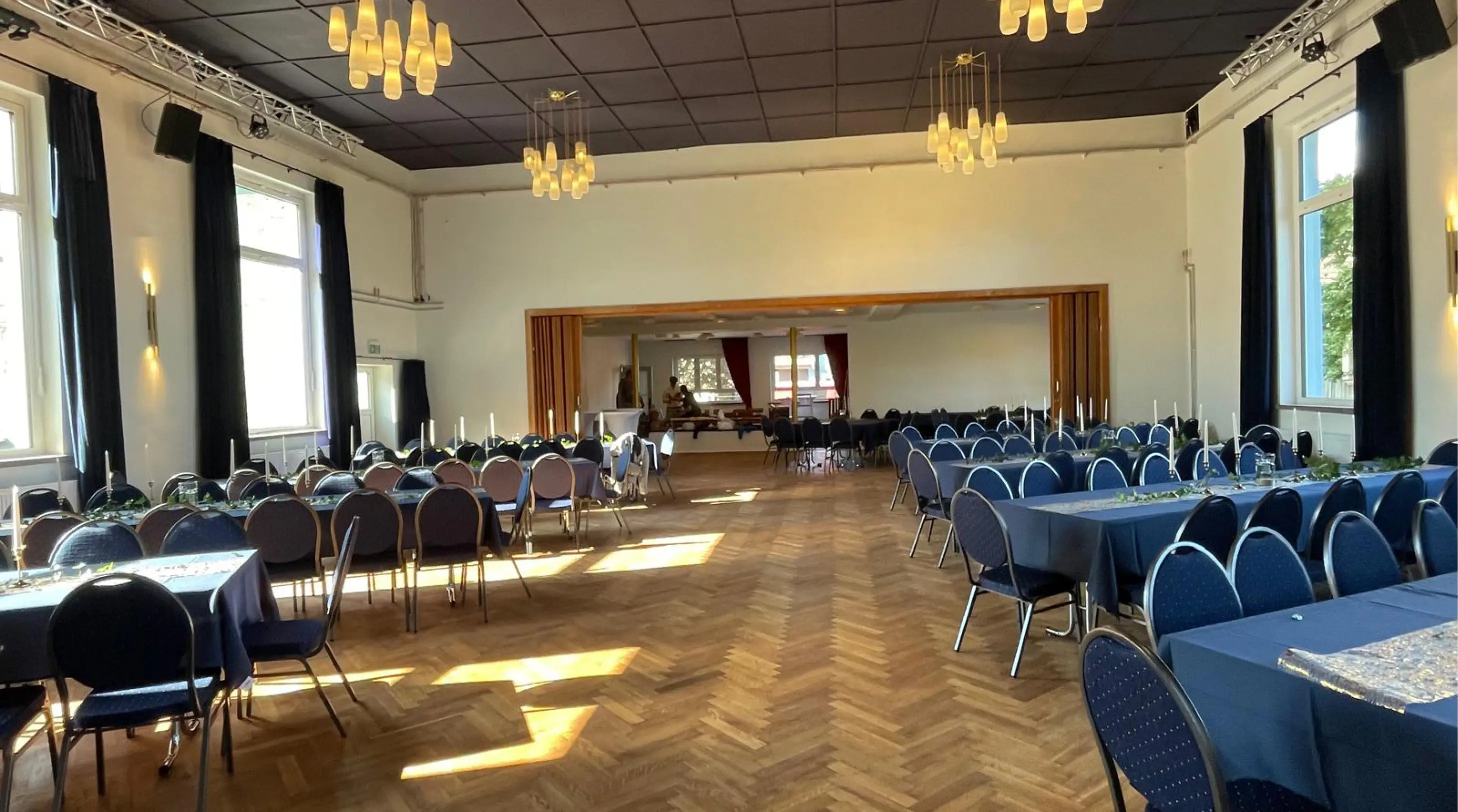 Großer Bankettsaal mit mehreren Tischen, die mit dunklen Tischdecken gedeckt und mit blauen Stühlen kombiniert sind, mit hohen Fenstern und Kronleuchtern.