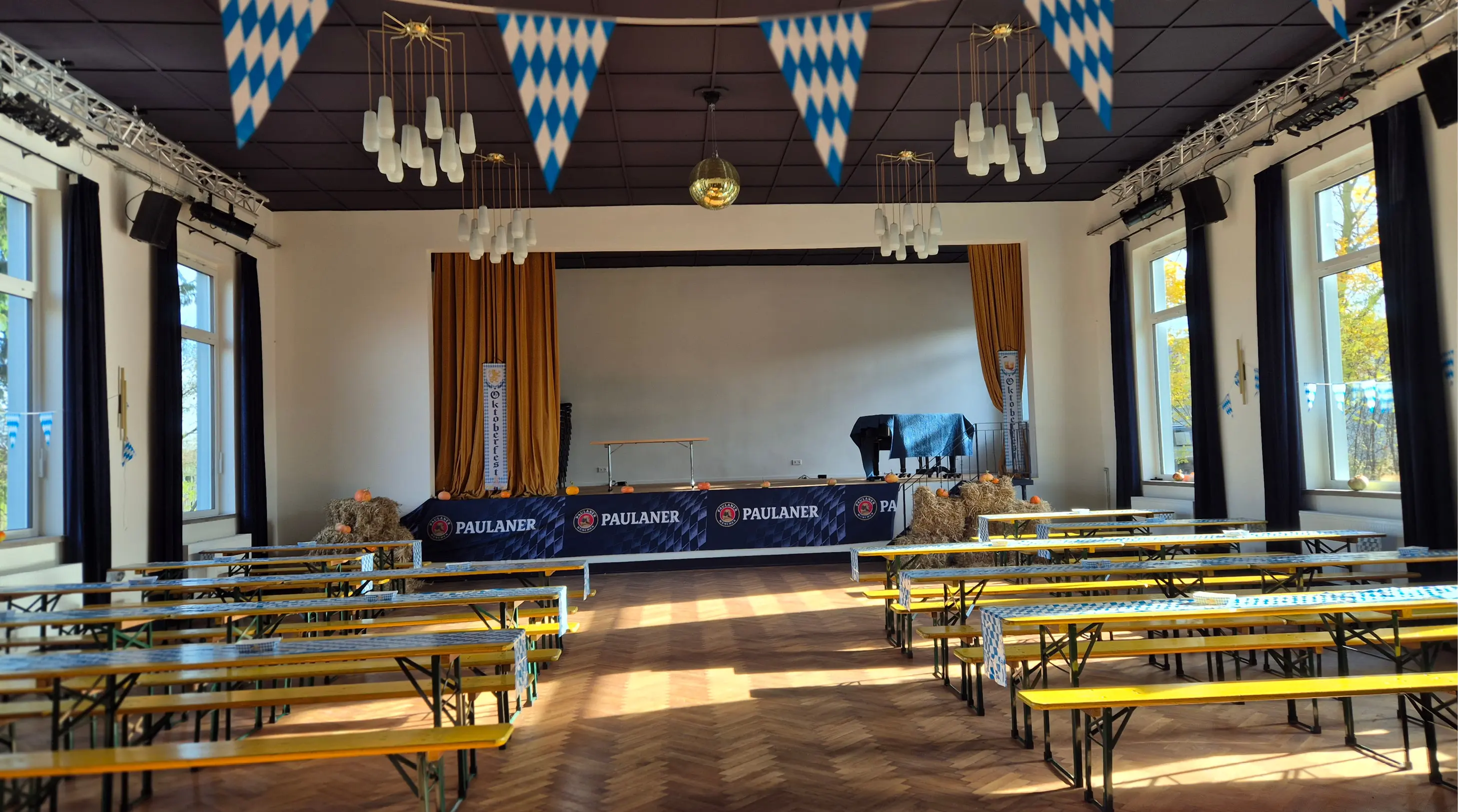 Helle Veranstaltungshalle mit gelben Bänken und Tischen, dekoriert für das Oktoberfest, mit blau-weißen Fahnen, Heuballen und einem Paulaner-Banner auf der Bühne.
