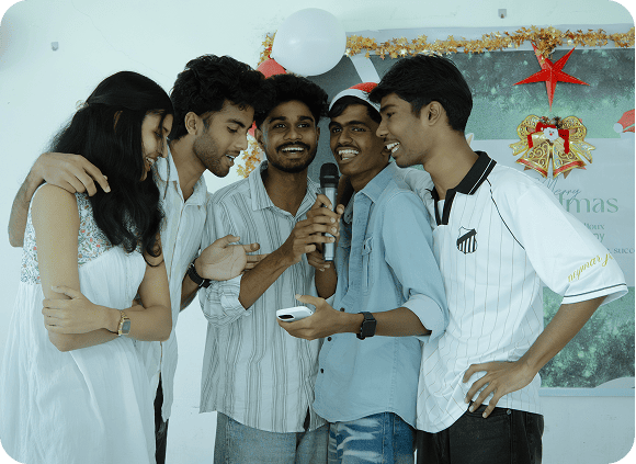 Five young adults singing together with a microphone at a festive indoor gathering.
