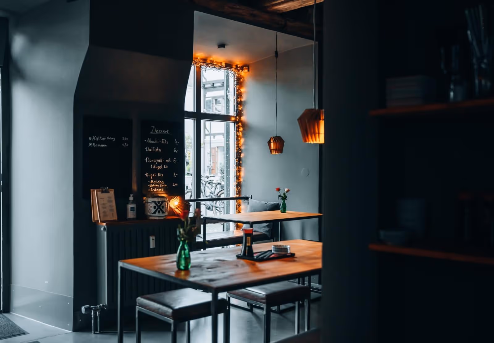 Innenansicht des Restaurants Yoko mit Holztischen, grünen Glasvasen, dekorativen Lampen und einer Desserttafel, gemütlich beleuchtet durch Lichterketten am Fenster.