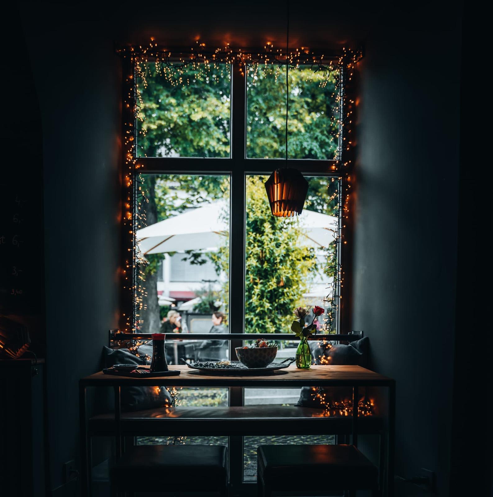Gemütlicher Sitzplatz im Restaurant Yoko am Fenster, dekoriert mit Lichterketten, Blick auf die Außenterrasse mit weißen Sonnenschirmen