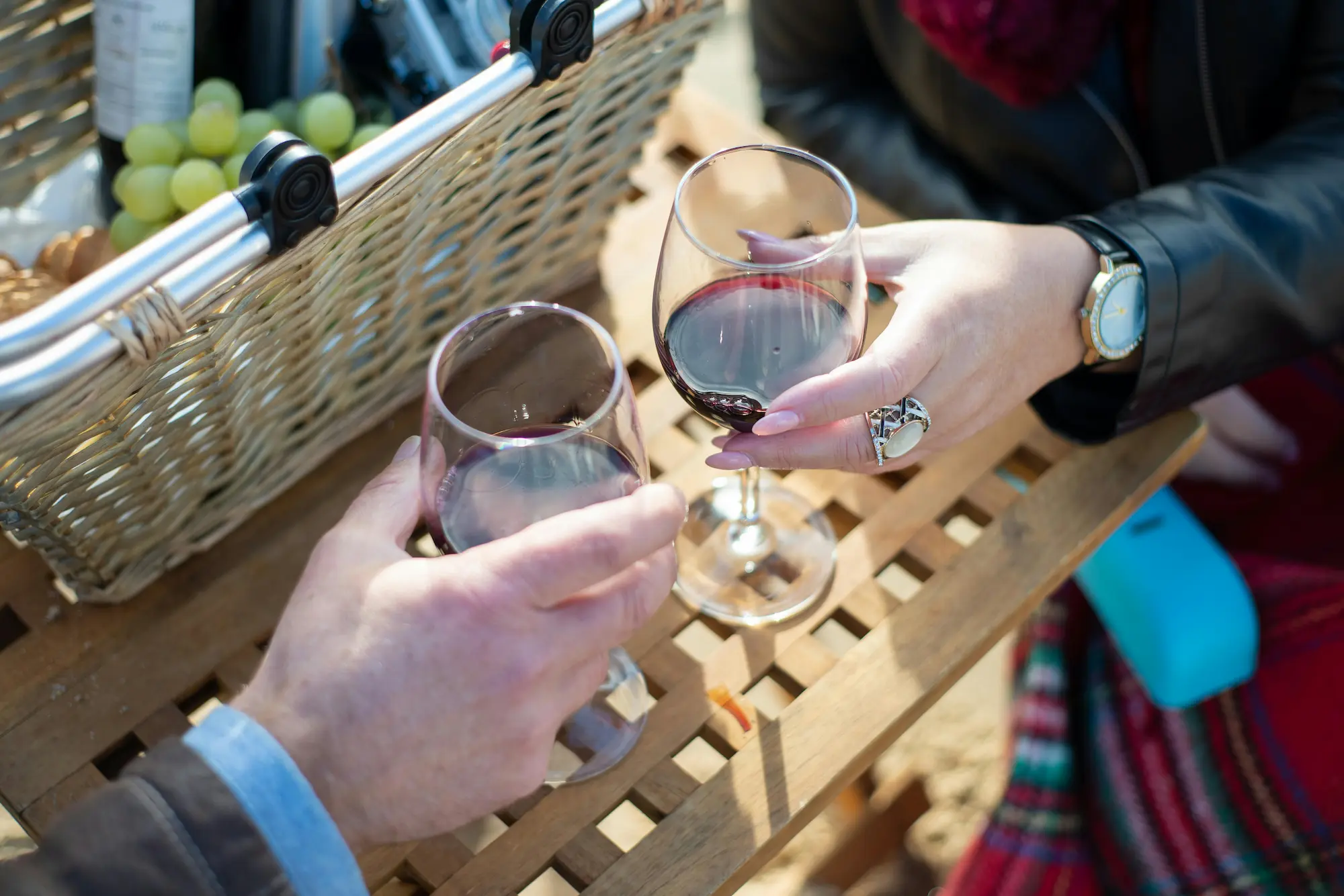 A couple toasts with wine glasses at a picnic table, enjoying a sunny day and each other's company.