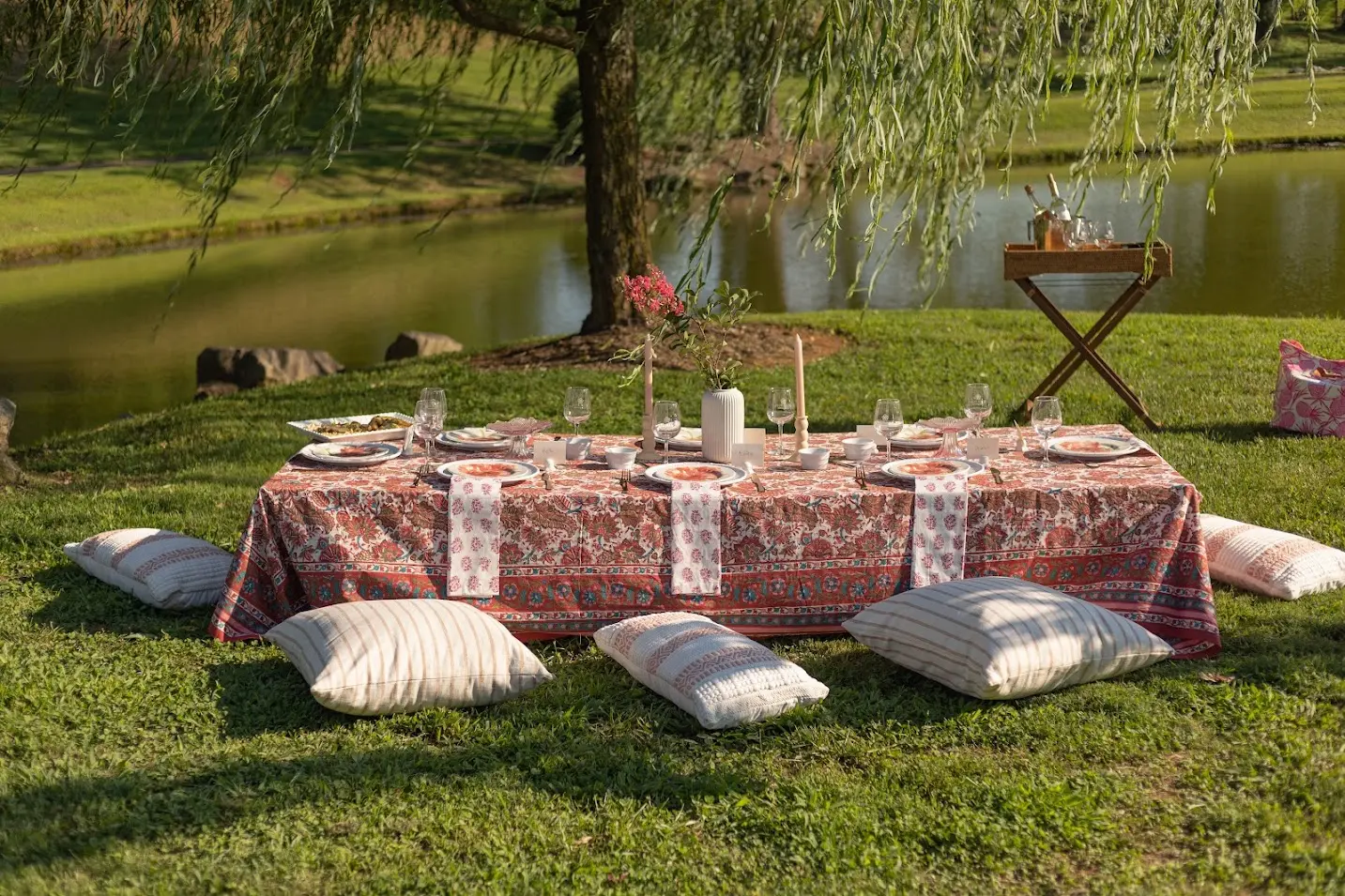Vineyard picnic setup with blush-patterned tablecloth, white cushions for seating, taper candles, and scenic pond with willow tree in the background.
