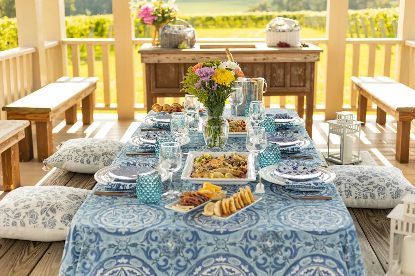 Beautiful vineyard pavilion picnic setup with a long table covered in a blue patterned tablecloth, elegant place settings, assorted appetizers, charcuterie, fresh flowers, and vineyard rows visible in the background.