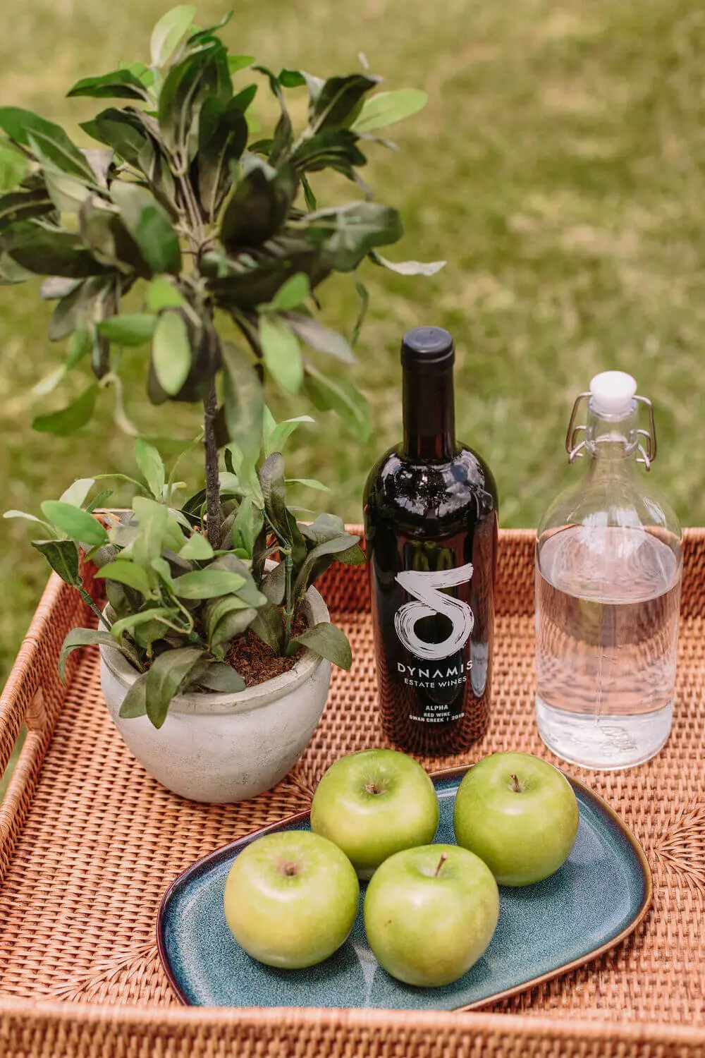 A basket containing green apples, a water bottle, and a small plant, arranged neatly on a simple surface.
