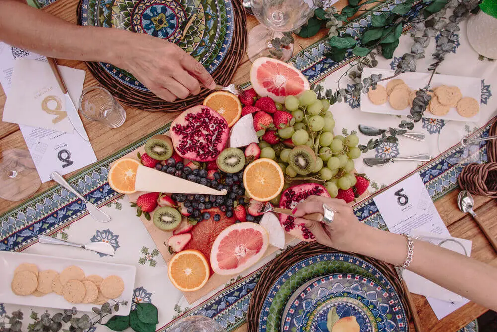 Beautifully arranged charcuterie board with flowers, fruit, and gourmet snacks on a colorful picnic setup.