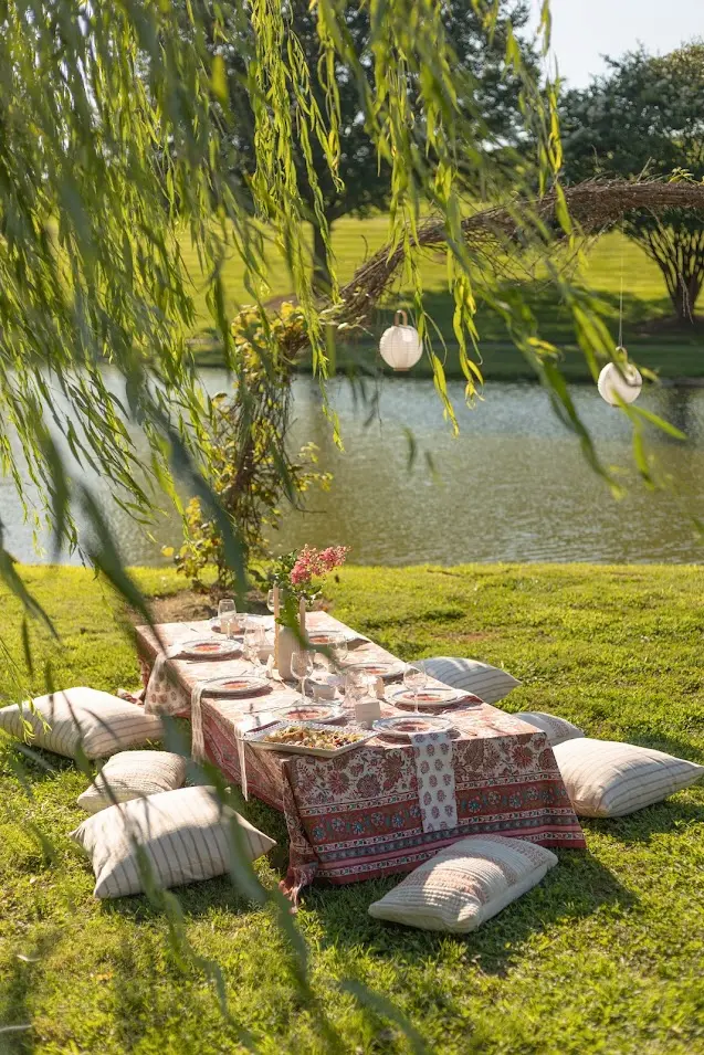 Low picnic table with white floral arrangements, wine glasses, and pillows under willow tree near pond.