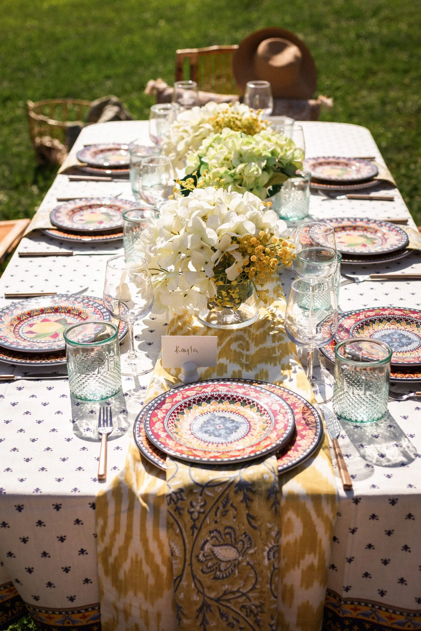 Outdoor vineyard picnic table with white and gold patterned tablecloth, pink and gold place settings, clear glassware, and white floral centerpieces under sunny skies.