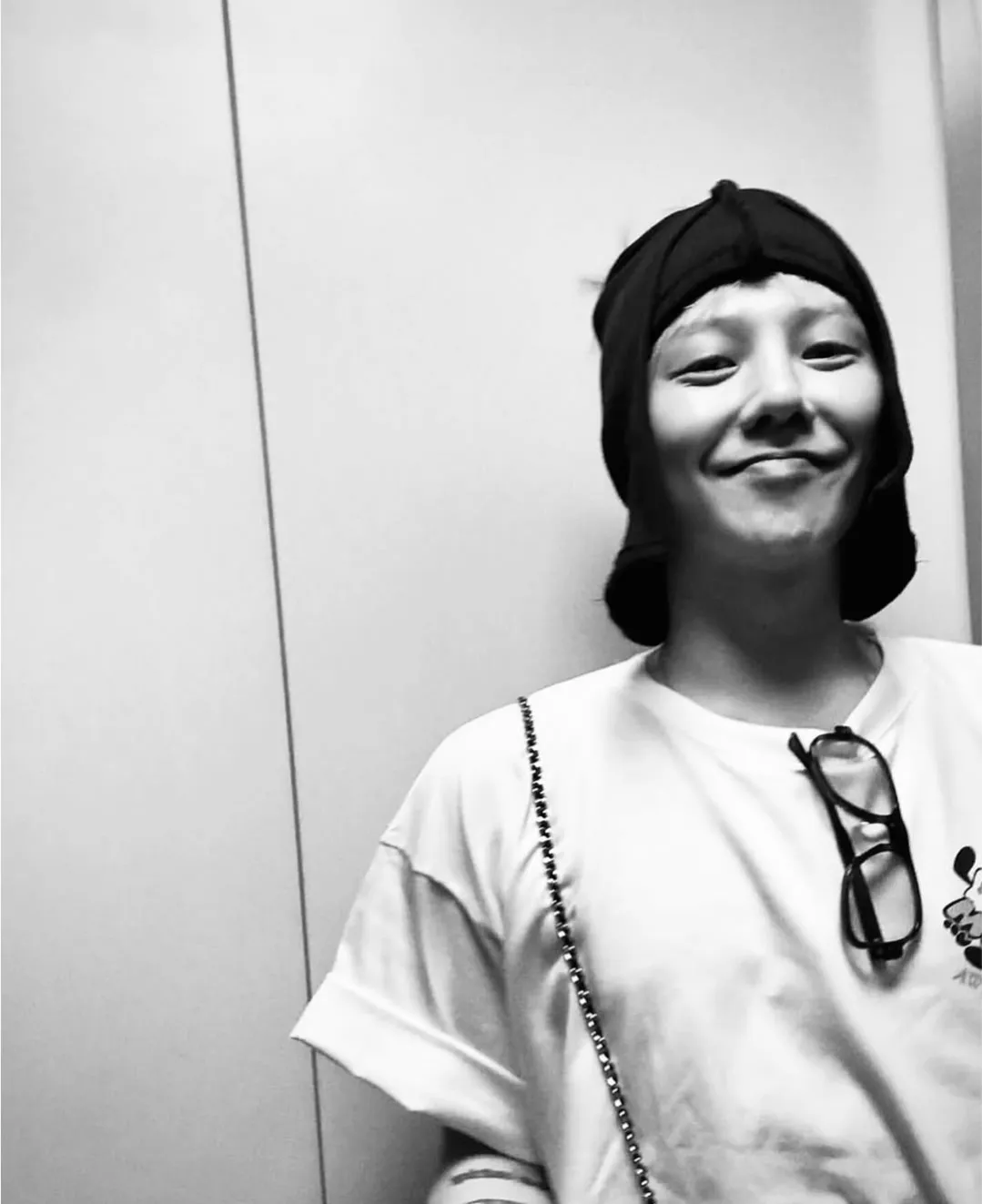 Black-and-white portrait of a person smiling casually, wearing a cap and a graphic T-shirt with glasses hanging from the neckline.