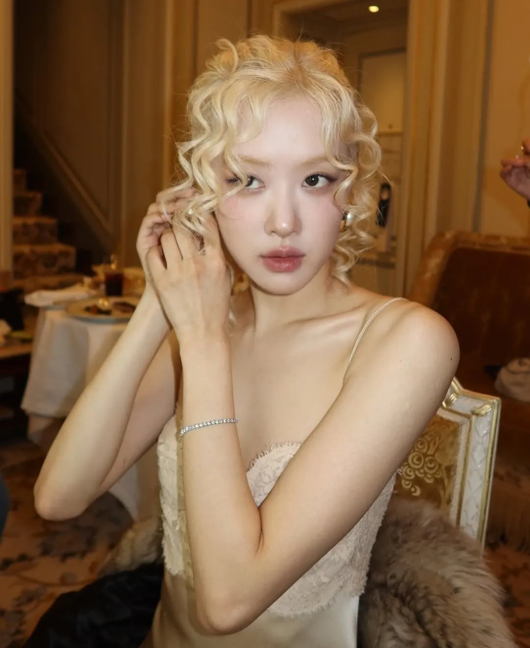 Person with curled blonde hair adjusting earrings in a luxurious interior setting.