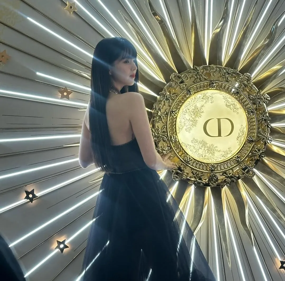 Kim Minju posing in a black backless dress in front of an ornate Dior backdrop featuring golden details and radiant light beams.
