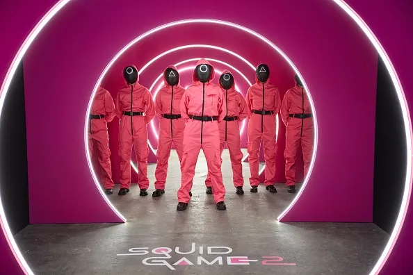 A group of guards in pink suits and black masks from Squid Game season 2 standing in a neon-lit circular hallway.