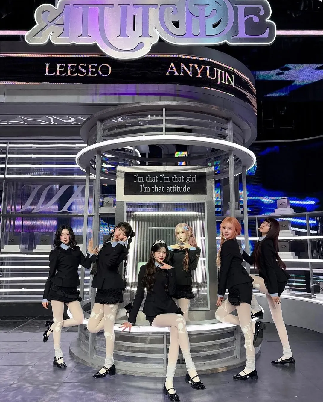 Six female K-pop idols pose confidently on a futuristic stage set with “attitude” signage and coordinated outfits.