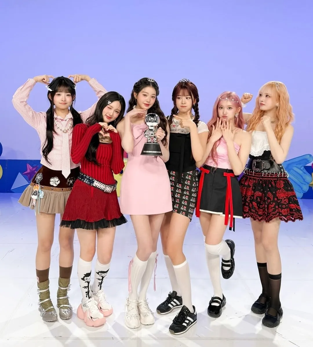 Six K-pop idols pose joyfully on stage holding a music show trophy, dressed in colourful and playful fashion styles.