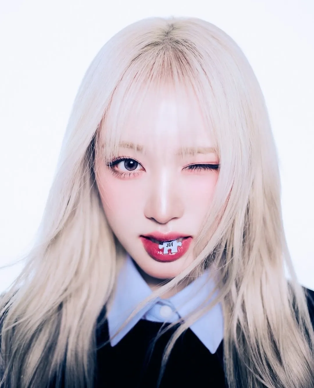 K-pop idol with platinum blonde hair winking while holding a tiny label with ‘JOY’ between her lips.