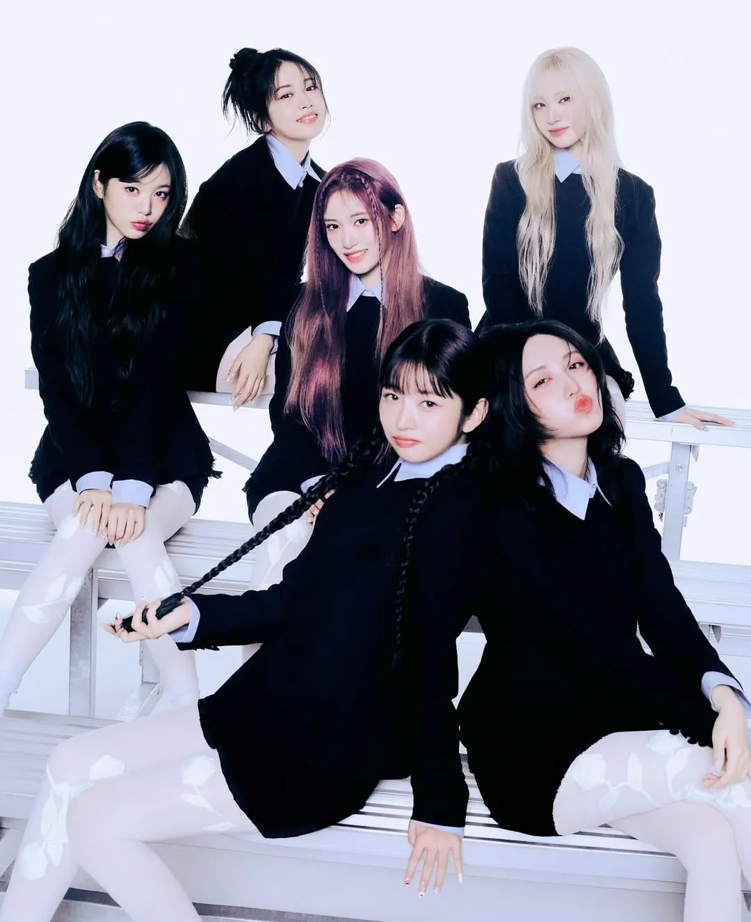 K-pop girl group poses in matching black school-uniform-inspired outfits with white tights against a clean white background.