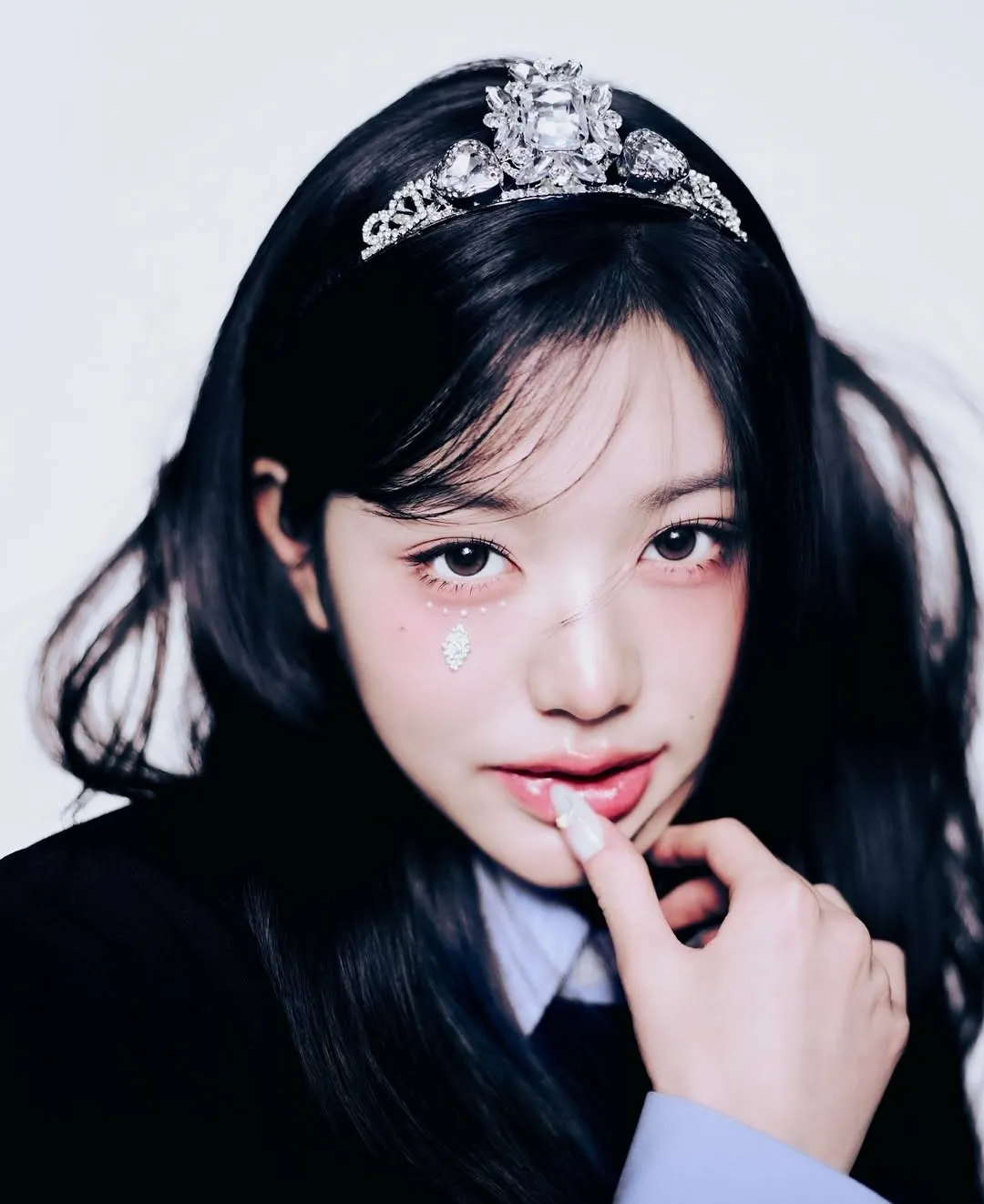Close-up of K-pop idol wearing a jeweled tiara and gem face decoration, softly posing with hand near lips.