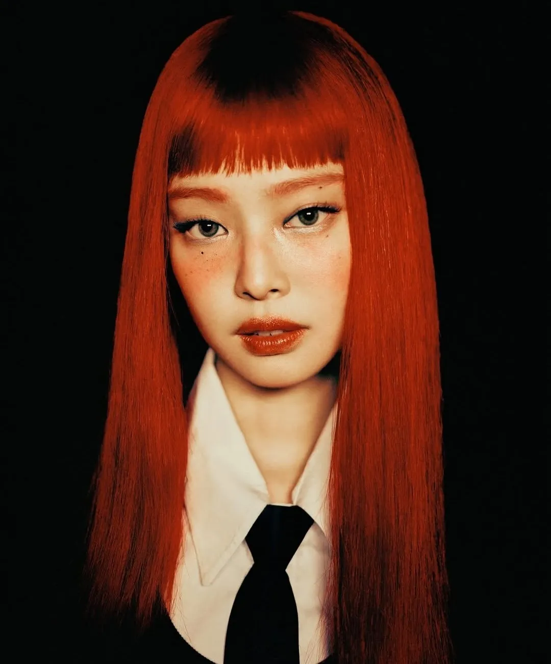Editorial portrait of a woman with straight red hair, bold makeup, and a black tie on a dark background.