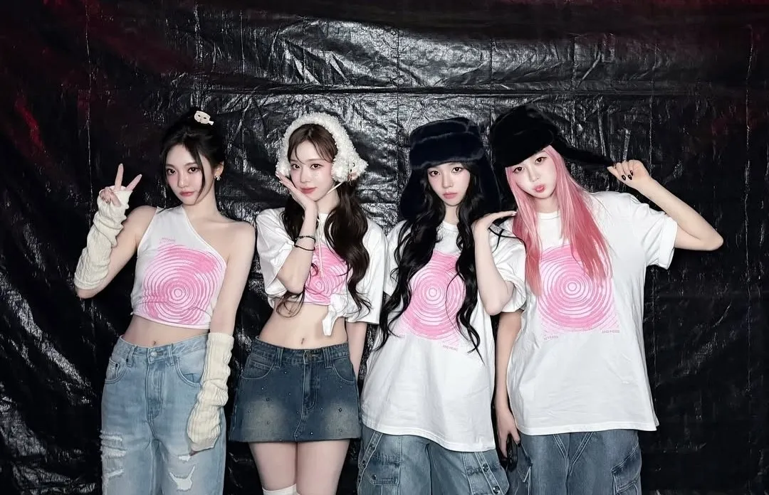 Four members of a girl group in denim and pink target print tops posing in front of a black backdrop.